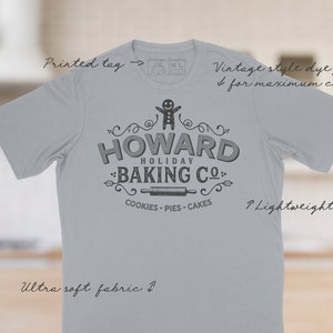May include: A light gray t-shirt with a vintage-style design featuring a gingerbread man and the text "HOWARD HOLIDAY BAKING CO." with the words "COOKIES - PIES - CAKES" underneath. The shirt is made of ultra-soft fabric and has a lightweight feel.