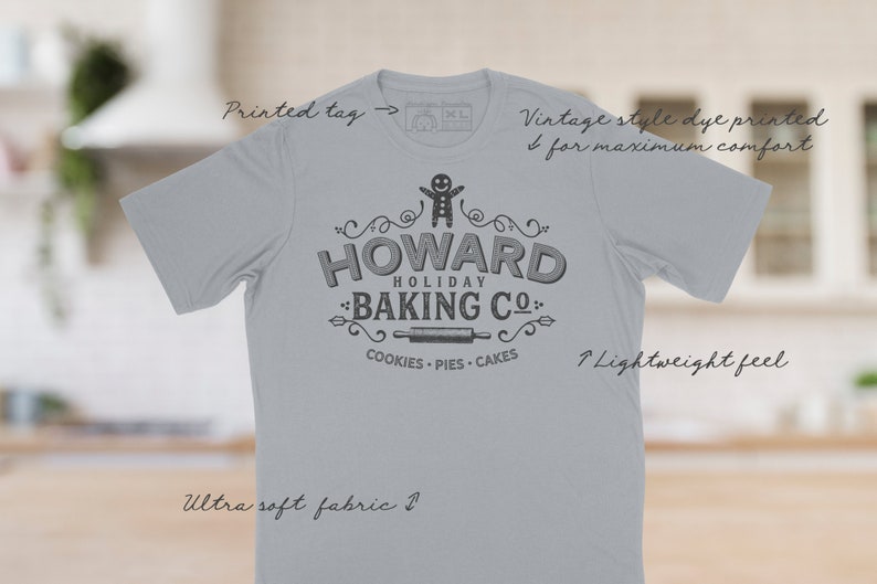 May include: A light gray t-shirt with a vintage-style design featuring a gingerbread man and the text "HOWARD HOLIDAY BAKING CO." with the words "COOKIES - PIES - CAKES" underneath. The shirt is made of ultra-soft fabric and has a lightweight feel.