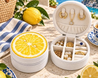 May include: A round white jewelry case with a lemon slice graphic on the lid. The open case reveals a beige interior with hooks for necklaces, a small pocket, and divided compartments containing various gold rings, pearl earrings, and a blue stone ring.