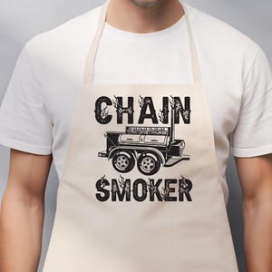 May include: A beige apron with the words "CHAIN SMOKER" in a bold, black font. The text is above and below a black illustration of a smoker grill. The apron is worn by a person wearing a white t-shirt.