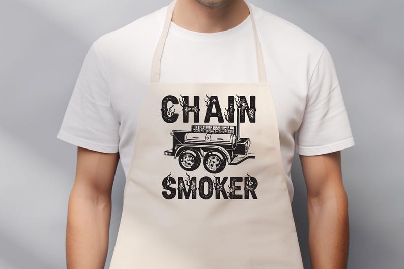 May include: A beige apron with the words "CHAIN SMOKER" in a bold, black font. The text is above and below a black illustration of a smoker grill. The apron is worn by a person wearing a white t-shirt.
