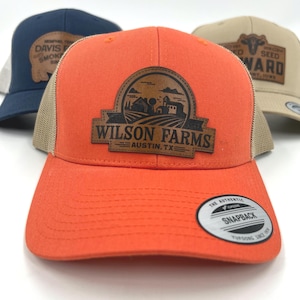 May include: Three trucker caps with curved peaks and mesh backs. The front cap is orange with a leather patch reading "WILSON FARMS AUSTIN, TX". The other caps are navy and tan with similar patches.