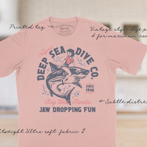 A pink t-shirt with a vintage style distressed print of a woman riding a shark with the text 'Deep Sea Dive Co. The Original Since 1940 Key West Florida Jaw Dropping Fun'.  These shirts are so buttery soft and comfy that you will want to wear it year round.  The designs and tags are dye printed for  comfort.   The shirts are available in many sizes and colors.
