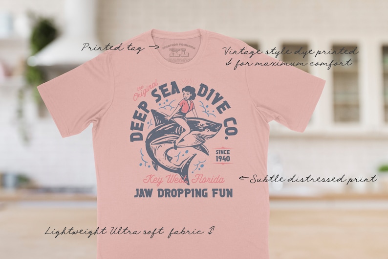 A pink t-shirt with a vintage style distressed print of a woman riding a shark with the text 'Deep Sea Dive Co. The Original Since 1940 Key West Florida Jaw Dropping Fun'.  These shirts are so buttery soft and comfy that you will want to wear it year round.  The designs and tags are dye printed for  comfort.   The shirts are available in many sizes and colors.