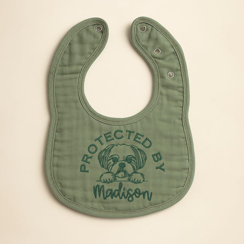 May include: Sage green baby bib with a rounded shape and snap closures. The bib features an embroidered design of a dog and the words "PROTECTED BY" above the name "Madison" in a matching green thread.