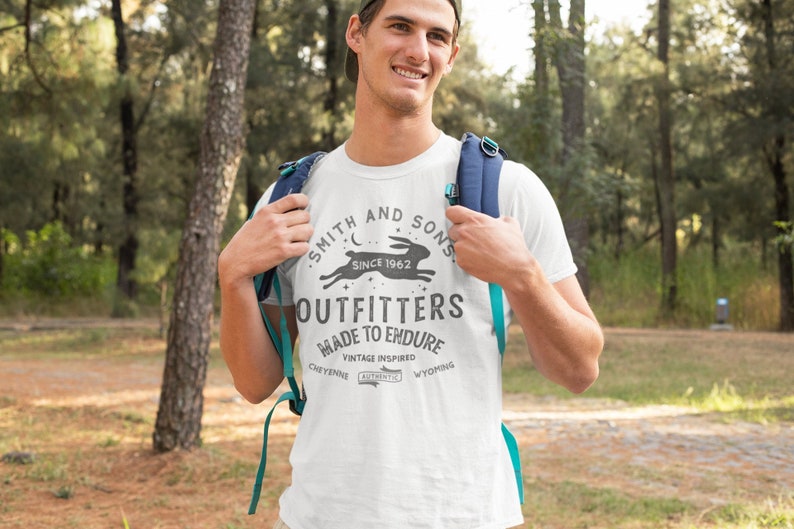 May include: White t-shirt with a vintage-inspired logo that reads "Smith and Sons Outfitters Since 1962 Made to Endure Vintage Inspired Cheyenne Authentic Wyoming". The logo features a running rabbit and a crescent moon.