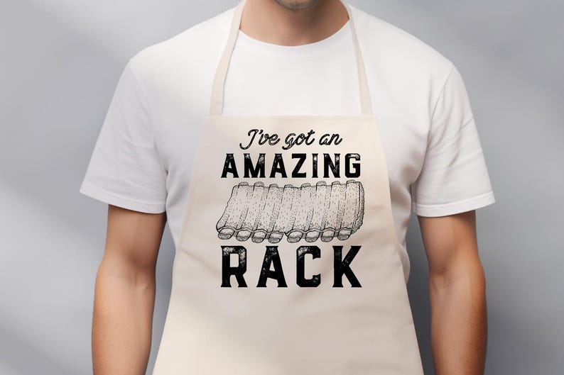 May include: Beige apron with the text "I've got an AMAZING RACK" in black. The design includes a graphic of a rack of ribs. The apron is worn by a person wearing a white t-shirt.