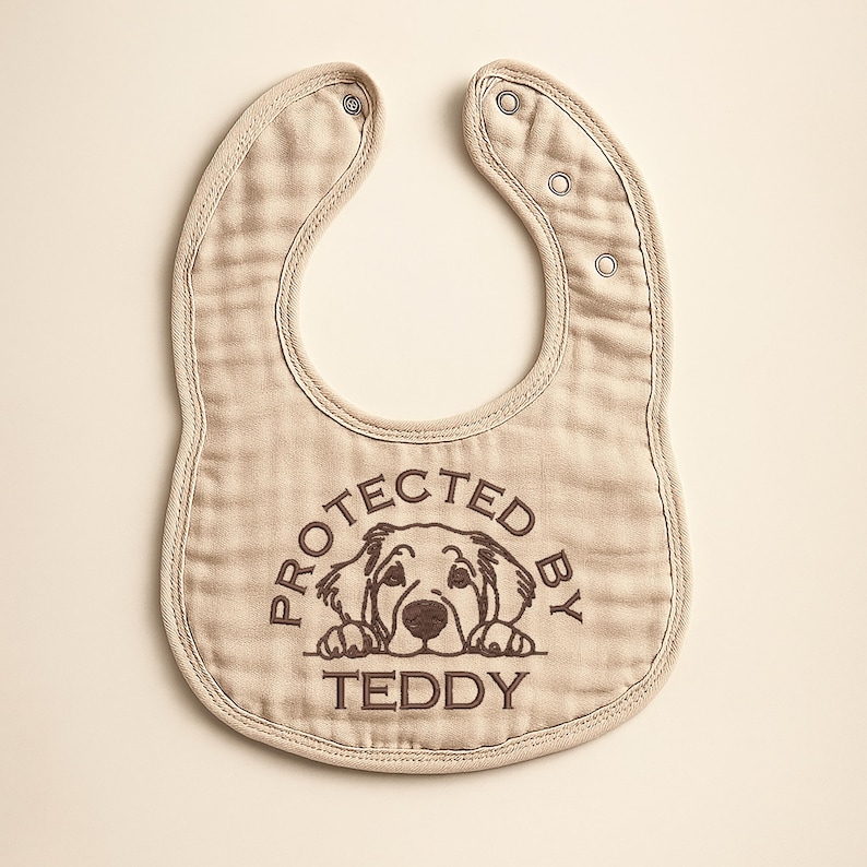 May include: A beige baby bib with a rounded shape and snap closures. The bib features a brown graphic of a dog and the text "PROTECTED BY TEDDY" in a circular design. The bib is made of a soft, textured fabric.