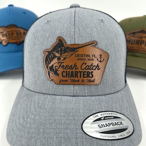 May include: Gray snapback hat with a black mesh back and a brown leather patch featuring a marlin illustration and the text "Fresh Catch Charters". The hat also has the text "Destin, FL Since 2015".