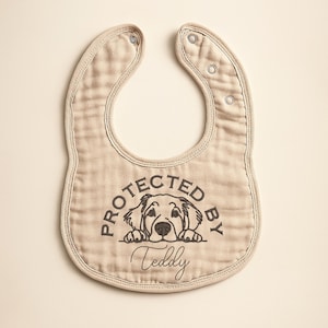 May include: Beige baby bib with a dog graphic and the text "PROTECTED BY Teddy". The bib has a rounded shape with snap closures at the neck. The bib is made of a soft, textured fabric.
