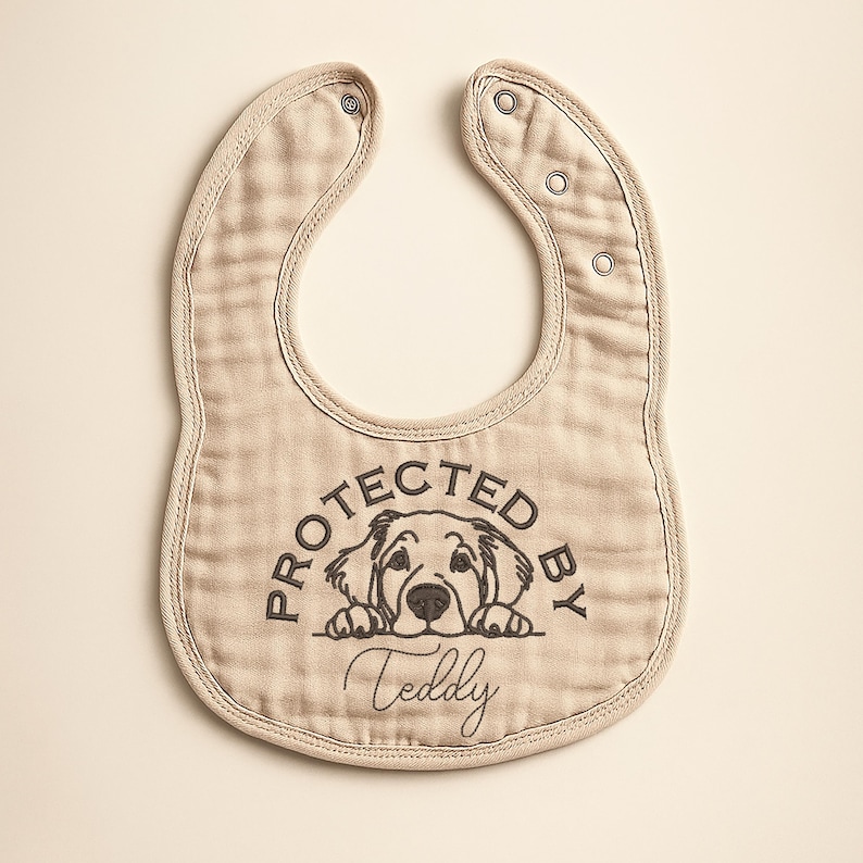May include: Beige baby bib with a dog graphic and the text "PROTECTED BY Teddy". The bib has a rounded shape with snap closures at the neck. The bib is made of a soft, textured fabric.