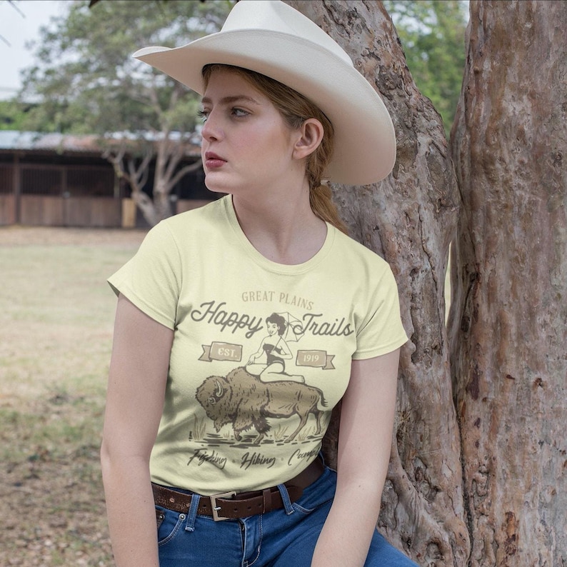 Saddle up for vintage vibes with our Happy Trails tee! Featuring a retro pinup cowgirl riding a mighty buffalo, this design blends classic Americana with bold Western charm. Distressed textures and warm, sun-faded tones give it that lived-in feel, perfect for road trips, festivals, or just kicking back in style. Soft, breathable fabric ensures all-day comfort on your wildest adventures. Ride off into the sunset with a smile!
