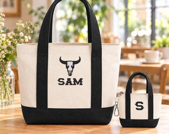 May include: A large off-white canvas tote bag with black handles and a black base, featuring an embroidered black bull skull and the text SAM. Beside it is a matching miniature canvas bag with a black base and the letter S.