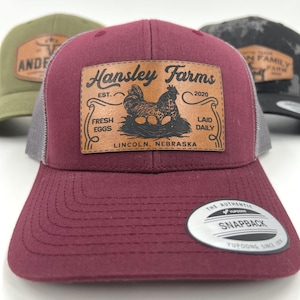 May include: A burgundy baseball cap with a gray mesh back and a curved brim. The cap features a leather patch with the text "Hansley Farms" and an illustration of a hen and eggs. The cap also has a snapback closure.