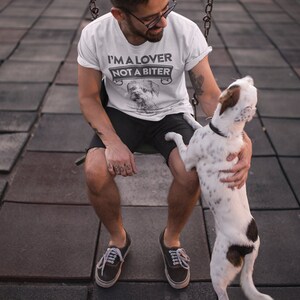 Op de afbeelding: Een wit T-shirt met de tekst "I'm a lover not a biter" en een klein hondenbeeld. De persoon die het shirt draagt, zit op een schommel met een hond.