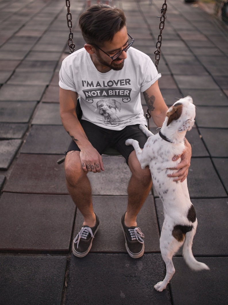 Op de afbeelding: Een wit T-shirt met de tekst "I'm a lover not a biter" en een klein hondenbeeld. De persoon die het shirt draagt, zit op een schommel met een hond.
