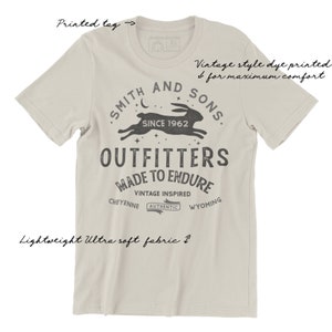 May include: A light beige t-shirt with a vintage-style print of a rabbit running with stars and the text "Smith and Sons, Since 1962, Outfitters, Made to Endure, Vintage Inspired, Cheyenne, Authentic, Wyoming".