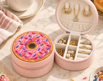 May include: A round, light pink jewelry box with a donut graphic on the lid featuring pink frosting and colorful sprinkles. The open interior reveals a cream-colored velvet lining with compartments for rings, necklaces, and earrings.