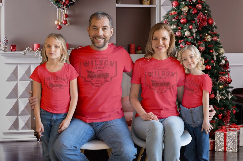 May include: A family of four wearing matching red t-shirts with a Christmas tree truck design and the text "Wilson Family Est. 2022". The family is standing in front of a decorated Christmas tree.