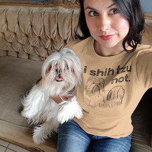 Op de afbeelding: Een vrouw draagt een beige t-shirt met een zwarte afbeelding van een Shih Tzu hond en de tekst "i shih tzu not." Ze zit op een bank met een witte Shih Tzu hond.