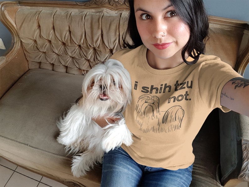 Op de afbeelding: Een vrouw draagt een beige t-shirt met een zwarte afbeelding van een Shih Tzu hond en de tekst "i shih tzu not." Ze zit op een bank met een witte Shih Tzu hond.