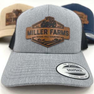 Op de afbeelding: Een grijze truckerpet met een zwarte mesh achterkant en een bruine leren patch. De patch toont de tekst "MILLER FARMS TEXAS HILL COUNTRY" met een tractorafbeelding. Een "YUPOONG" snapback-tag is zichtbaar op de klep.