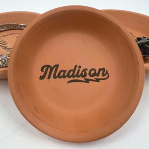 Puede incluir: Tres platos de arcilla de terracota. El plato central presenta el nombre "Madison" en una fuente de escritura negra. Los platos son redondos con forma de cuenco poco profundo, perfectos para guardar joyas o objetos pequeños. Los platos son de color naranja cálido y terroso.
