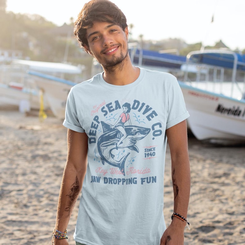 Make a splash with this bold Deep Sea Dive Co. tee featuring a vintage pin-up girl riding a fierce shark like a retro sea queen. Inspired by old-school tattoo flash and classic dive aesthetics, this shirt brings serious nautical attitude with a wink. Perfect for lovers of the deep blue, beachcombers, and retro enthusiasts who aren’t afraid to swim with sharks. Dive in and wear it loud!