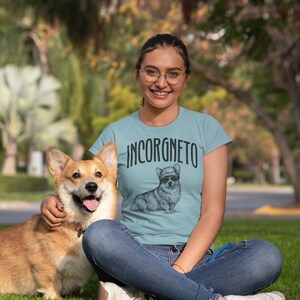 Op de afbeelding: Een vrouw draagt een lichtblauw T-shirt met een zwart-witte illustratie van een corgi die een zonnebril en een hoed draagt. De tekst "INCORGNETO" staat op het shirt gedrukt. Ze zit op het gras met een corgi-hond.