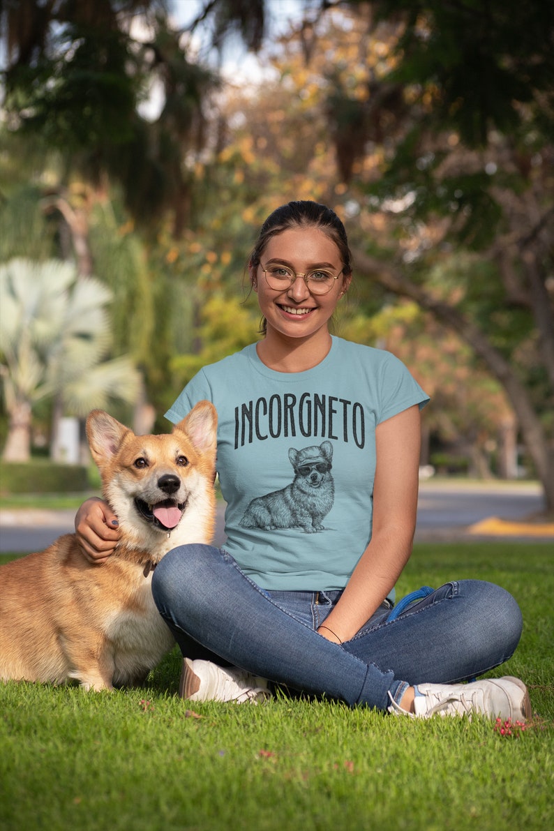 Op de afbeelding: Een vrouw draagt een lichtblauw T-shirt met een zwart-witte illustratie van een corgi die een zonnebril en een hoed draagt. De tekst "INCORGNETO" staat op het shirt gedrukt. Ze zit op het gras met een corgi-hond.