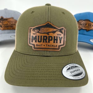 Może przedstawiać: Zielona czapka typu trucker z brązową skórzaną naszywką z ilustracją ryby i napisem "MURPHY BAIT & TACKLE". Czapka ma zakrzywiony daszek i zapięcie na zatrzask. Tył jest z beżowej siatki.