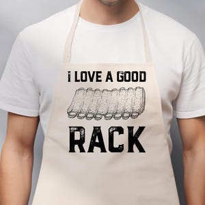 May include: Beige apron with the text "I LOVE A GOOD RACK" in black. The apron features a graphic of a rack of ribs. The apron is worn by a person wearing a white t-shirt.