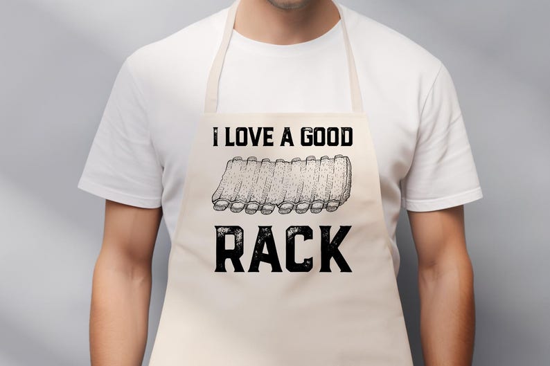 May include: Beige apron with the text "I LOVE A GOOD RACK" in black. The apron features a graphic of a rack of ribs. The apron is worn by a person wearing a white t-shirt.