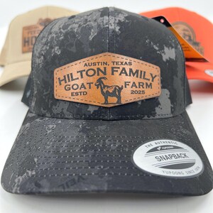 May include: Black and gray camouflage-patterned snapback hat with a leather patch that reads "HILTON FAMILY GOAT FARM ESTD 2025" and features a goat illustration. The hat has a curved brim and a snapback closure.