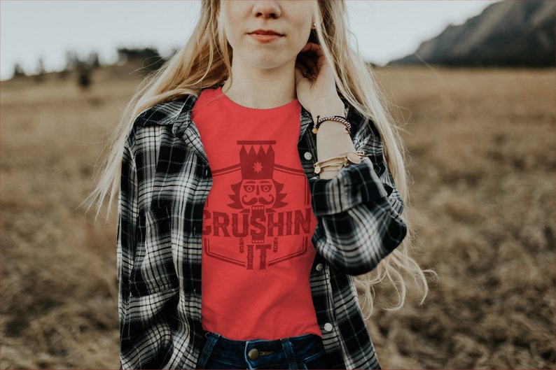 May include: A red t-shirt with a graphic of a nutcracker with a crown and the text "CRUSHING IT" in a vintage style font.