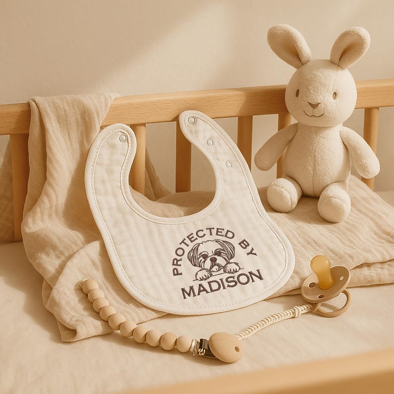 May include: A baby bib with the text "PROTECTED BY MADISON" and a dog graphic. A plush bunny, pacifier, and wooden bead clip are also present. The items are arranged on a crib.