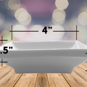 May include: A white rectangular ceramic dish, 10 cm long, 6.35 cm wide, and 2.54 cm deep. The dish has a simple, modern design with slightly flared sides. It is suitable for serving appetizers or small portions.