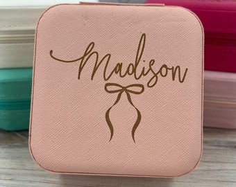 May include: A pink jewelry box with a gold inscription that reads "Madison" with a gold bow underneath the name.