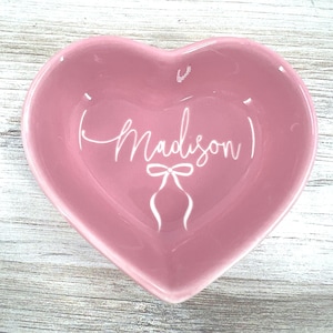 May include: A pink heart-shaped ceramic trinket dish with the name "Madison" written in white cursive lettering. A white bow is also written on the dish.