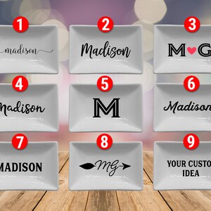 May include: Nine white rectangular ceramic dishes with various black designs. These include names, initials, and custom text. The dishes are presented on a wooden surface, offering a range of personalised options.