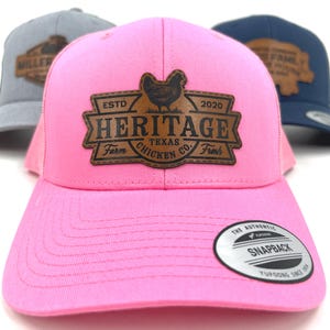 May include: Pink Heritage Farm Texas Chicken Co. hat with a brown leather patch featuring a chicken graphic. The hat has a curved brim and a snapback closure. Additional hats in gray and navy blue are visible in the background.