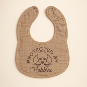 May include: A tan baby bib with a rounded shape and snap closures. The bib features the embroidered text "PROTECTED BY Pebbles" with a poodle illustration. The bib is made of soft fabric and has a stitched border.
