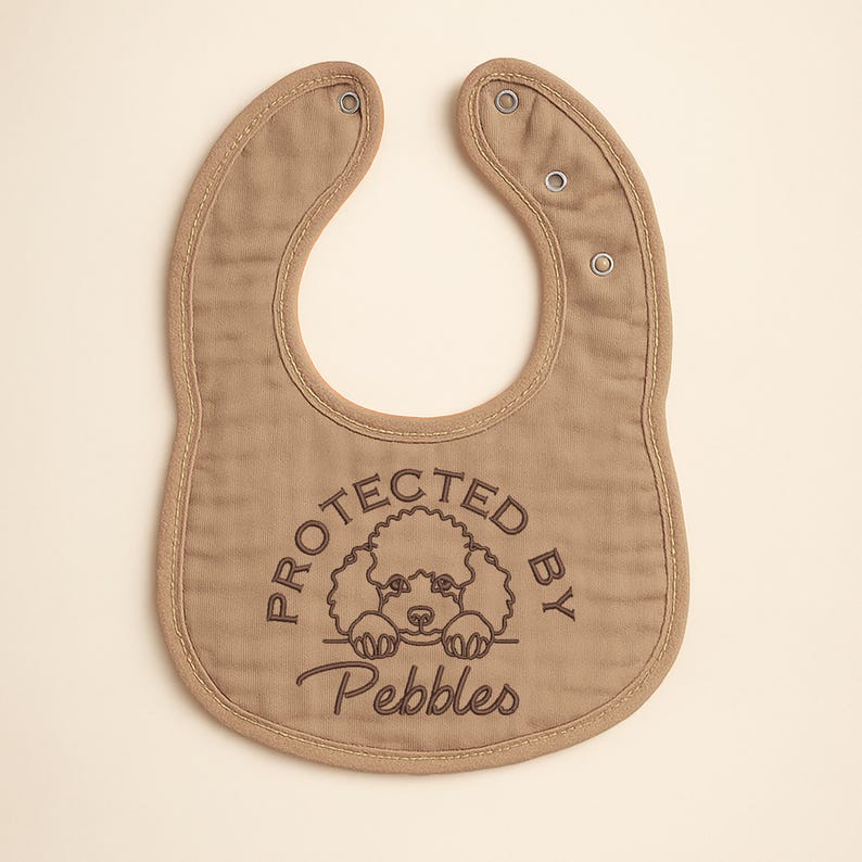 May include: A tan baby bib with a rounded shape and snap closures. The bib features the embroidered text "PROTECTED BY Pebbles" with a poodle illustration. The bib is made of soft fabric and has a stitched border.