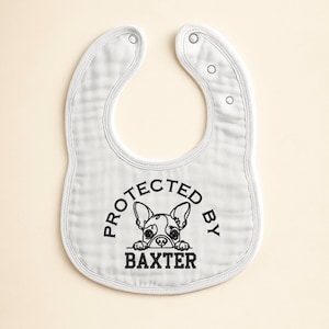 May include: White baby bib with a rounded shape and snap closures. The bib features the text "PROTECTED BY BAXTER" in black, with a graphic of a French Bulldog. The bib is made of a soft, textured fabric.