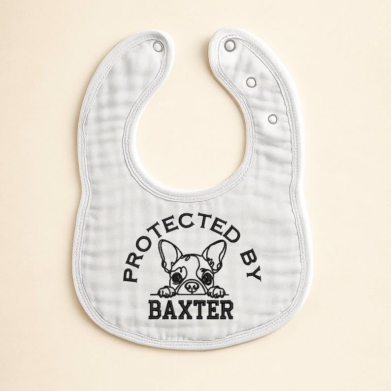 May include: White baby bib with a rounded shape and snap closures. The bib features the text "PROTECTED BY BAXTER" in black, with a graphic of a French Bulldog. The bib is made of a soft, textured fabric.