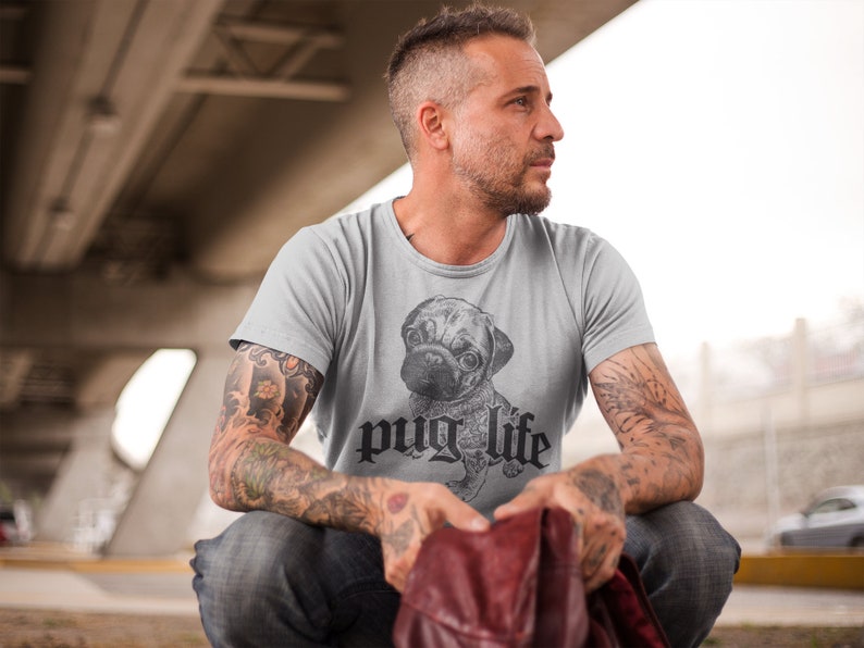 Op de afbeelding: Een grijs T-shirt met een zwart-witte illustratie van een mopshond en de tekst "pug life" in een gotisch lettertype.