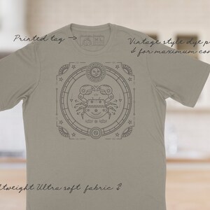 Puede incluir: Una camiseta gris claro con un dise&ntilde;o de impresi&oacute;n de tinte de estilo vintage que presenta un s&iacute;mbolo del zod&iacute;aco de cangrejo en un c&iacute;rculo con estrellas y otros s&iacute;mbolos. La camiseta tiene una etiqueta impresa que dice "XL".