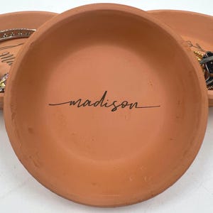 May include: A terracotta-coloured, shallow dish with the name "madison" in black script. The dish is round and appears handmade. Other dishes are visible in the background, some with jewellery.