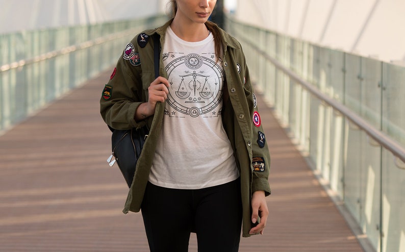 May include: A woman wearing a green jacket with patches and a white t-shirt with a black and white graphic design of a scale. She is wearing black pants and carrying a black backpack.