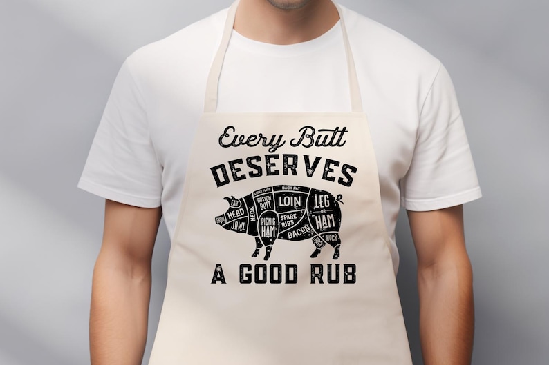May include: A beige apron with the text "Every Butt Deserves A Good Rub" and a diagram of a pig with cuts of meat labeled. The apron is worn by a person wearing a white t-shirt.